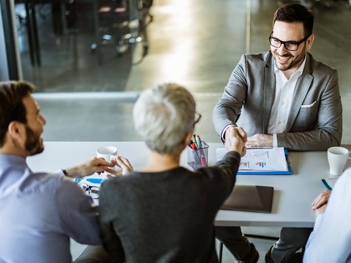 Geschäftstreffen mit vier Personen, ein Mann im Anzug lächelt und gibt einer Person über den Tisch hinweg die Hand.