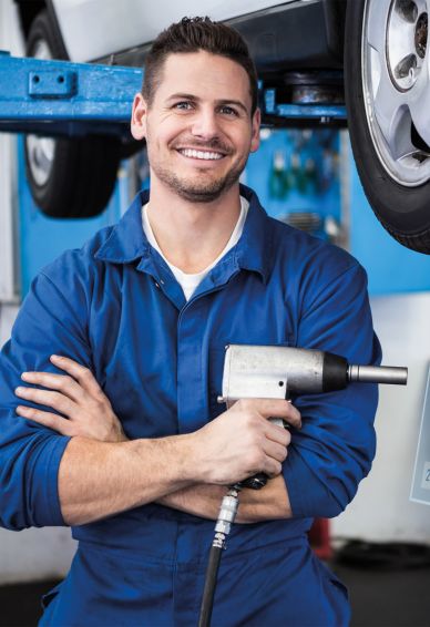 Kfz-Mechaniker in blauer Arbeitskleidung hält eine Schraubpistole und lächelt in einer Werkstatt vor einem angehobenen Auto.